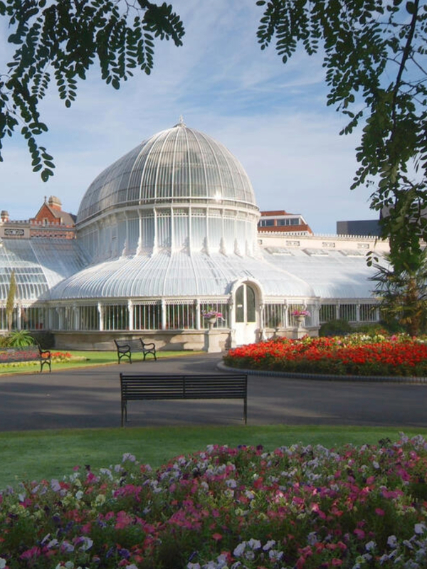 Victorian domed glasshouse in Belfast Botanic Gardens, framed by flowerbeds and benches on a bright day.