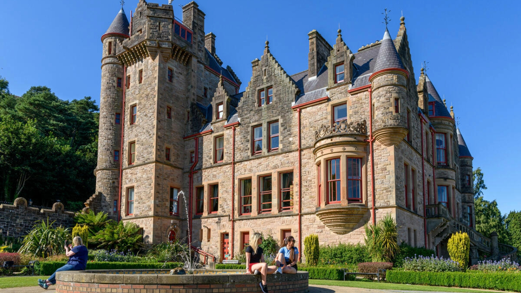 Die steinernen Türme von Belfast Castle überblicken die gepflegten Gärten, in denen sich Besucher an sonnigen Tagen an einem Springbrunnen entspannen können.