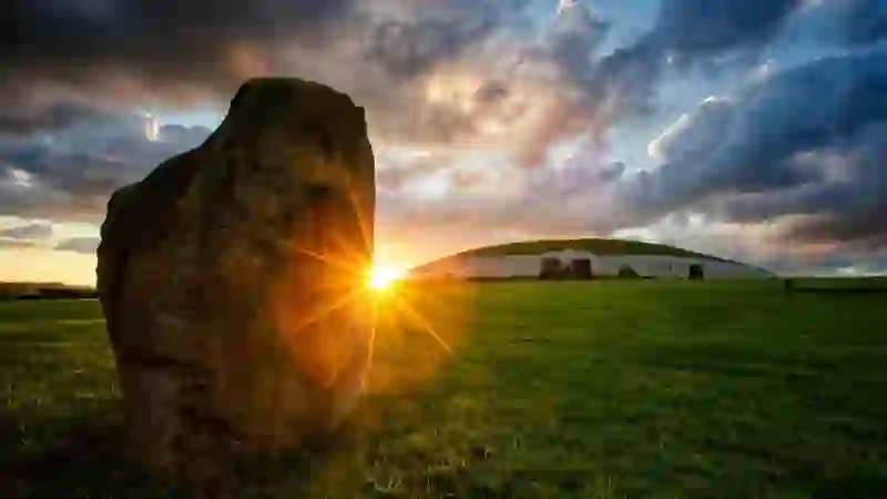 Standing stone at sunrise facing Newgrange passage tomb, County Meath, beneath dramatic clouds.