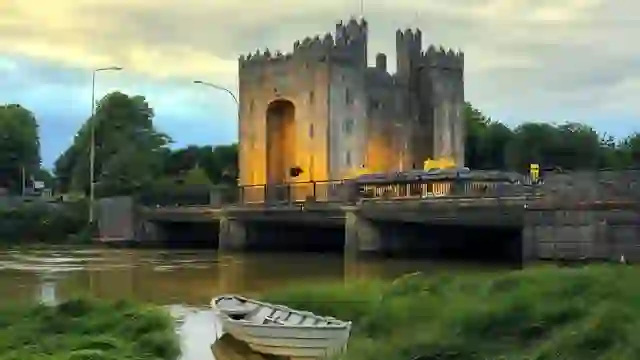 Bunratty Castle illuminated beside the River Ratty at dusk, with a small boat resting in the reeds in the foreground.