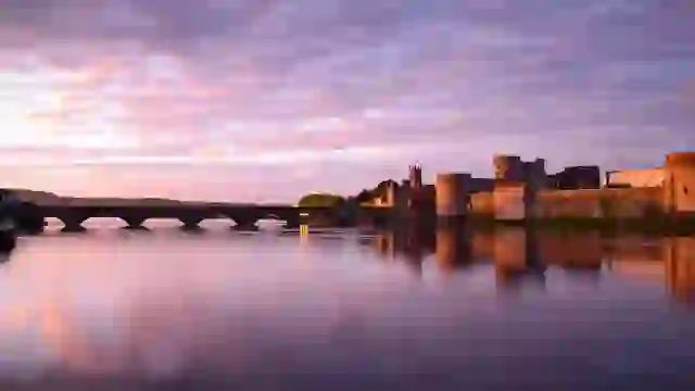 King John’s Castle in Limerick reflected in the River Shannon at sunset, with a stone bridge stretching across the water.