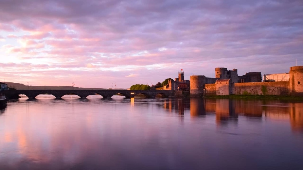 King John’s Castle in Limerick reflected in the River Shannon at sunset, with a stone bridge stretching across the water.