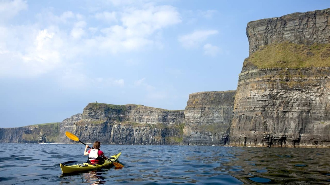 Kayaker paddling beneath the Cliffs of Moher on the Atlantic Ocean, with towering limestone cliffs in the background.