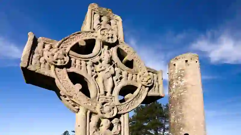 Close-up van een gebeeldhouwd hoog kruis in Clonmacnoise, met op de achtergrond een ronde toren, in county Offaly.