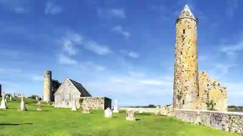 Kloosternederzetting Clonmacnoise met ronde torens, kerkruïne en grafstenen onder een blauwe hemel in county Offaly.