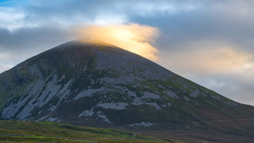 Der Berg Croagh Patrick bei Sonnenuntergang, mit goldenem Licht, das durch die Wolken über dem Gipfel in der Grafschaft Mayo, Irland, bricht.