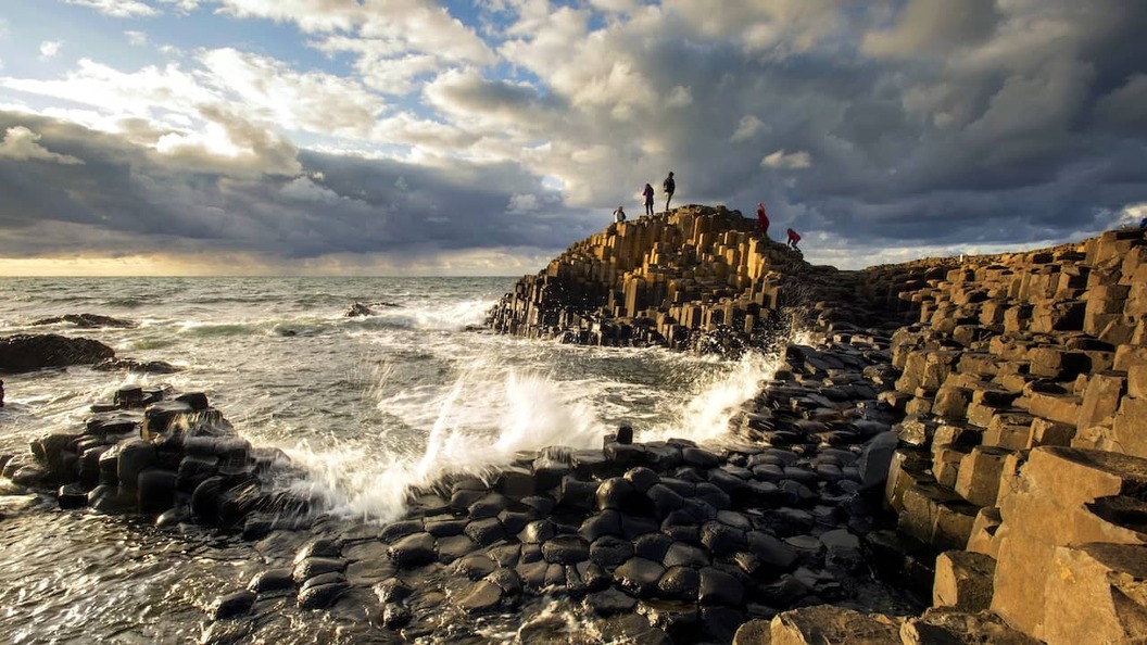 Visitors exploring the Giant's Causeway's basalt columns as Atlantic waves crash beneath dramatic skies.