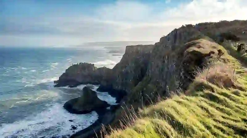 Walkers on a grassy clifftop path overlooking dramatic sea cliffs and waves along the Causeway Coast.