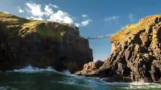 Carrick-a-Rede Rope Bridge spanning dramatic sea cliffs in County Antrim, with waves crashing below under blue sky.