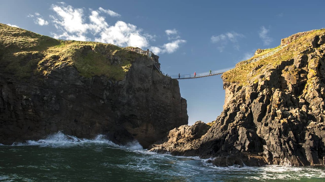Carrick-a-Rede Rope Bridge spanning dramatic sea cliffs in County Antrim, with waves crashing below under blue sky.