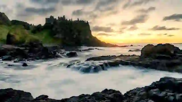 Dunluce Castle ruins perched on a cliff at sunset above waves crashing against the Antrim shoreline.