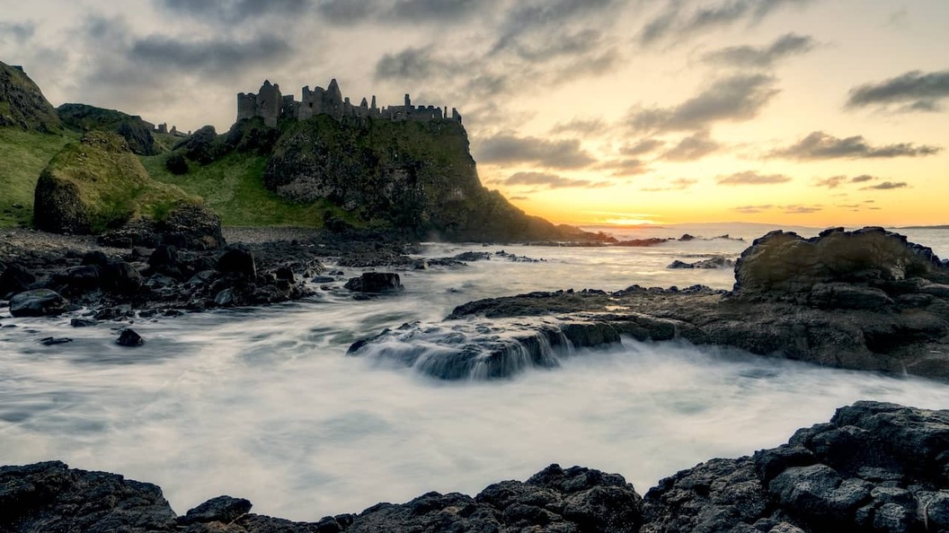 Dunluce Castle ruins perched on a cliff at sunset above waves crashing against the Antrim shoreline.