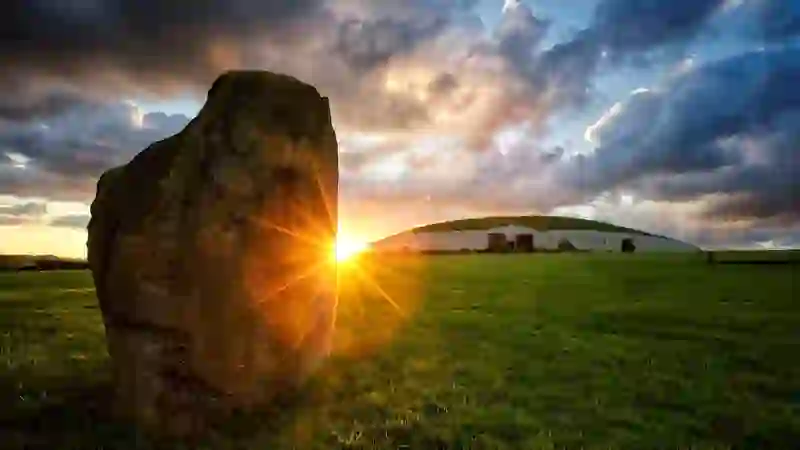 Large standing stone and Newgrange mound at sunrise, with golden light breaking through dramatic clouds.