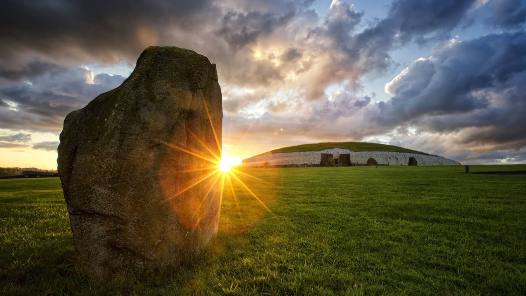 Large standing stone and Newgrange mound at sunrise, with golden light breaking through dramatic clouds.