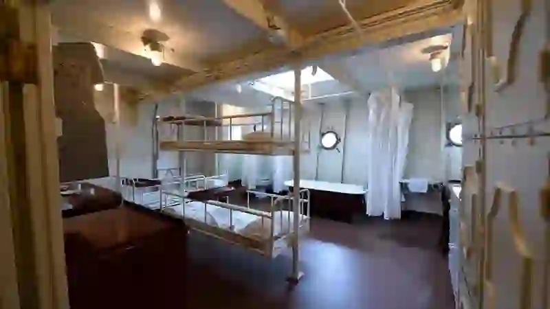 Schlafquartiere auf der HMS Caroline mit Metallstockbetten, einem Waschbereich mit Vorhängen und Bullaugenfenstern.