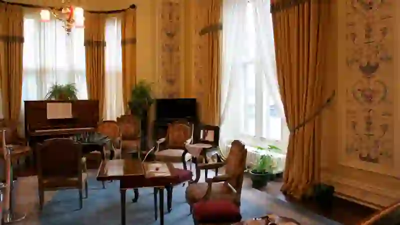 Elegante sala vittoriana all'interno dell'Abbazia di Kylemore con pianoforte, tavolo da scacchi e alte finestre drappeggiate.