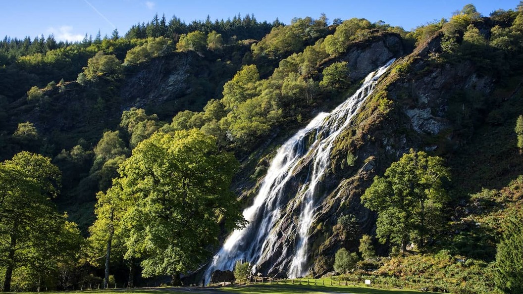 Powerscourt Waterfall cascading down a rocky, forested hillside, surrounded by lush green trees beneath a clear blue sky.