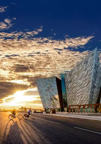 Cyclists passing Titanic Belfast at sunset, with golden light illuminating the building’s striking exterior.