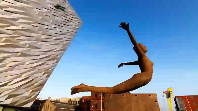 Rostskulptur einer ins Wasser tauchenden Figur neben der eckigen Fassade des Titanic Belfast unter einem klaren blauen Himmel.