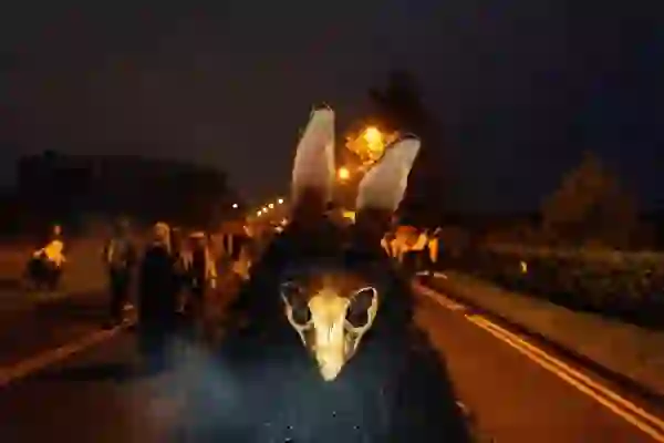 Person in a dark cloak wearing a horned animal skull mask with tall ears, standing on a road at night during the Púca Festival.