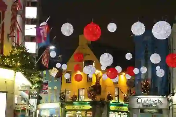 Festive street scene in Galway with glowing red and white Christmas baubles hanging above colourful shopfronts decorated with lights and greenery.