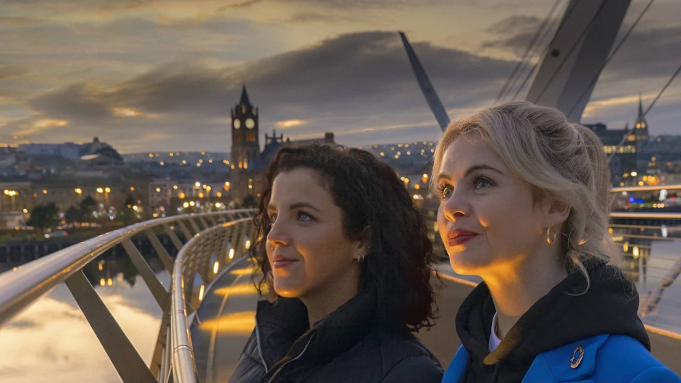 Due donne ammirano il paesaggio urbano dal Peace Bridge di Derry~Londonderry al tramonto, con il Guildhall e le luci della città che brillano alle spalle.