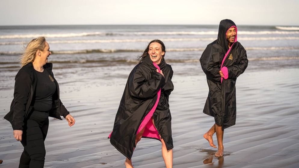 Amigos envueltos en cómodas batas de baño riéndose juntos en una playa después de un baño en el mar con Mussenden Unwind en el condado de Londonderry, con suaves olas de fondo.