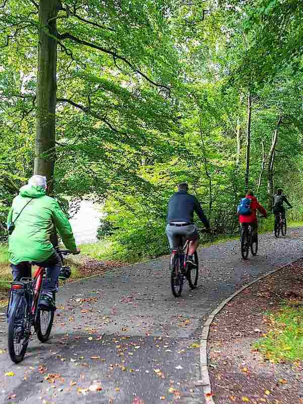 Cyclists exploring a leafy greenway through Derry~Londonderry, surrounded by woodland and riverside scenery.