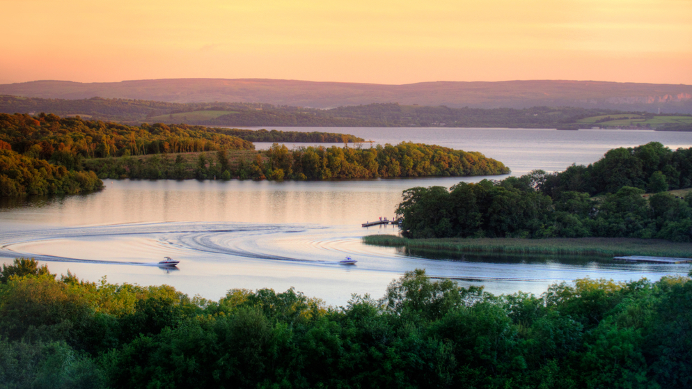 Motorboats create sweeping wakes across Lough Erne at sunset, surrounded by wooded islands in County Fermanagh.