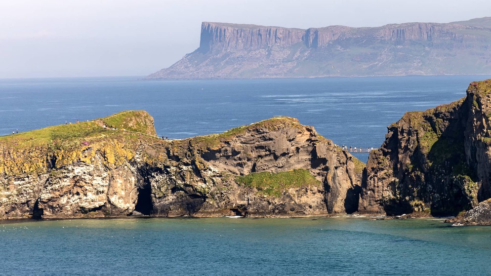 Clifftop views of Carrick-a-Rede with the rope bridge visible above turquoise water and distant headlands in County Antrim.