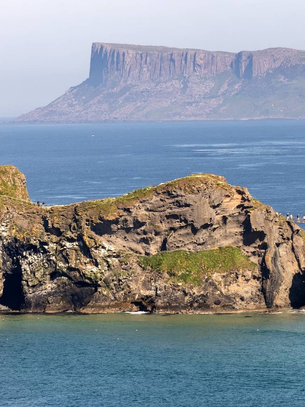 Clifftop views of Carrick-a-Rede with the rope bridge visible above turquoise water and distant headlands in County Antrim.