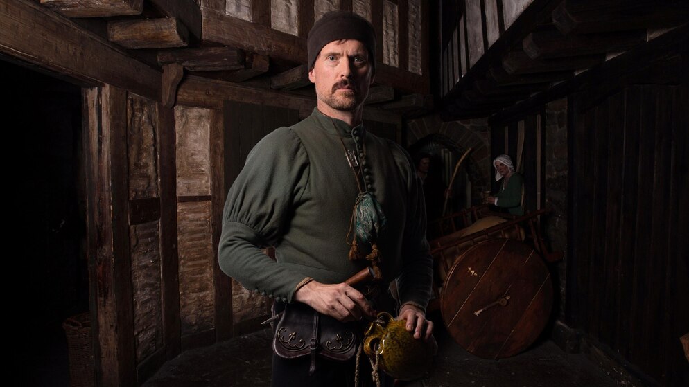 Man dressed as a Viking with green tunic and cap inside a recreated timber building in Ireland.