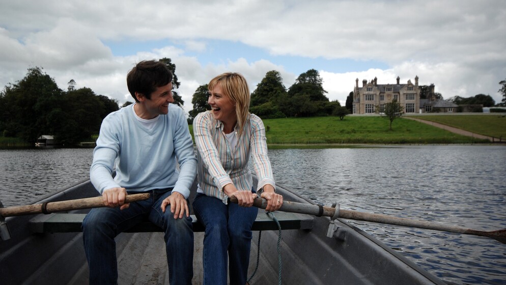 Couple laughing in a rowboat on a lake with a stately home and lush green grounds behind, promoting wedding adventures.