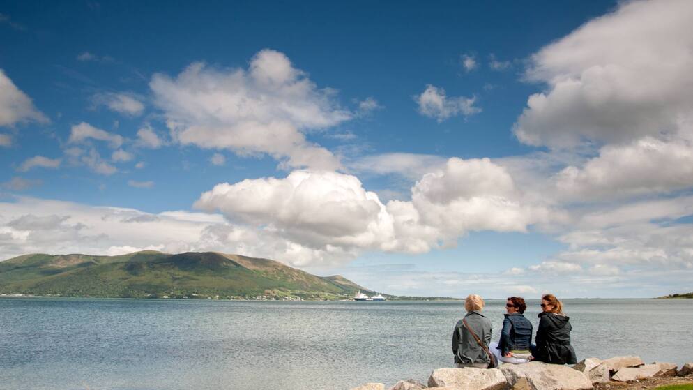 Three women walking on the coast with mountains and blue sky in the background.