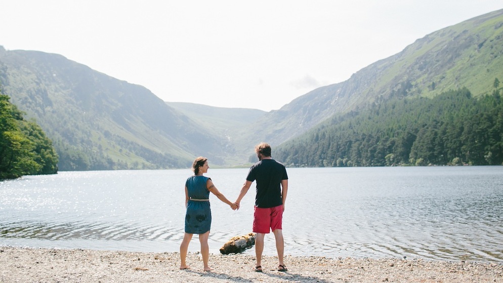 Koppel hand in hand bij een meer in Glendalough, county Wicklow, omlijst door beboste bergen onder een bleke hemel.
