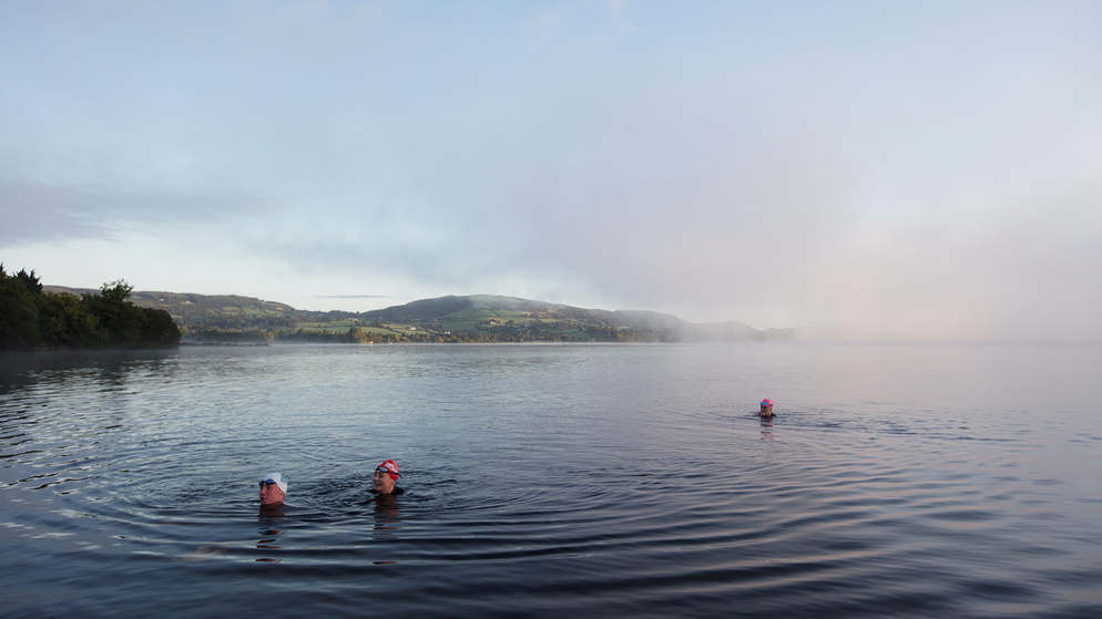 Two swimmers in misty lake water with pink hats near treeline under pale sky at dawn.