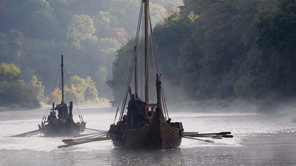 Nachbildung eines Wikinger-Langschiffs, das durch Morgennebel segelt, mit bewaldeten Hügeln im Hintergrund.