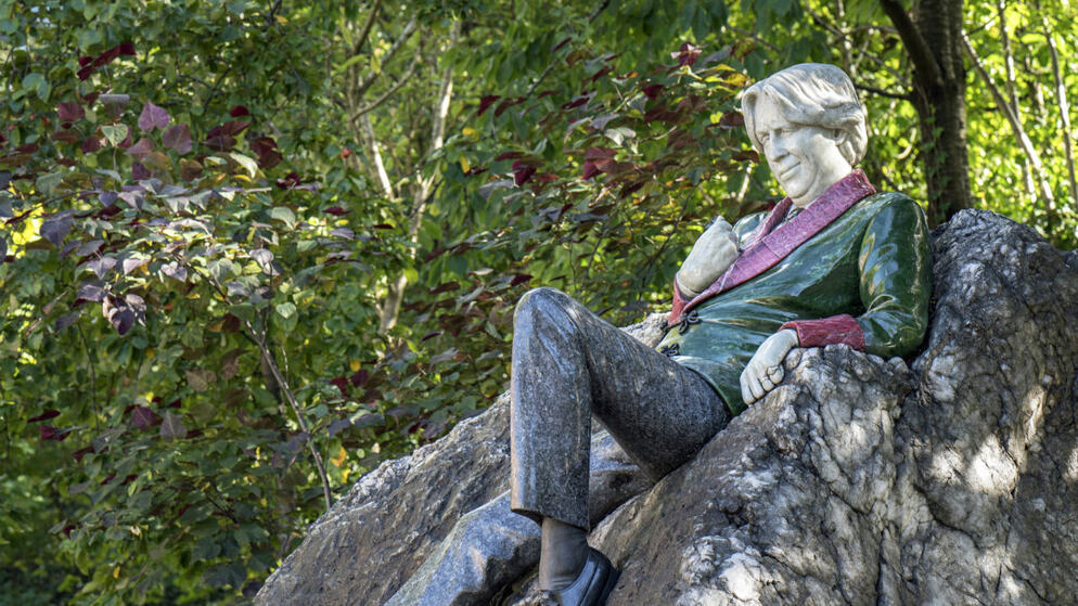Bunte Statue von Oscar Wilde, der sich auf einem Felsen im Merrion Square entspannt, umgeben von blühenden Bäumen in Dublin.