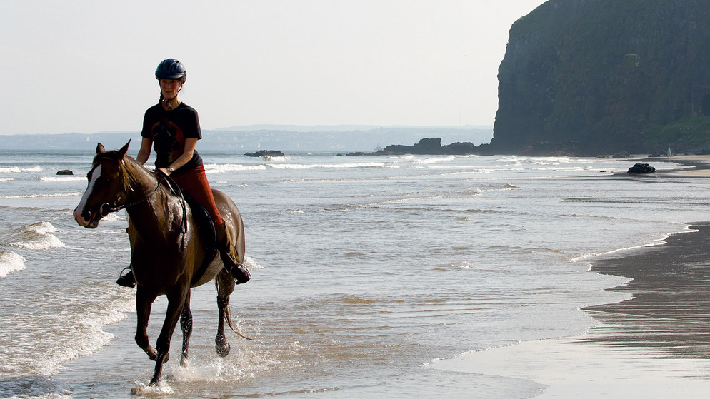 Horse rider along a sandy beach in Ireland with waves rolling in and cliffs in the background.