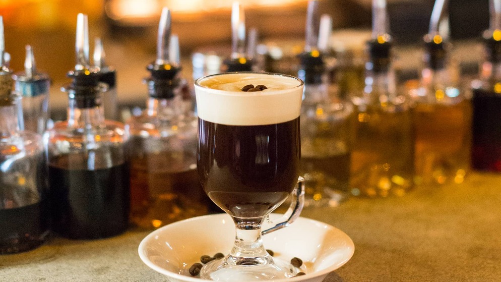 Glass of Irish coffee topped with cream and coffee beans, served against a background of liqueur bottles.