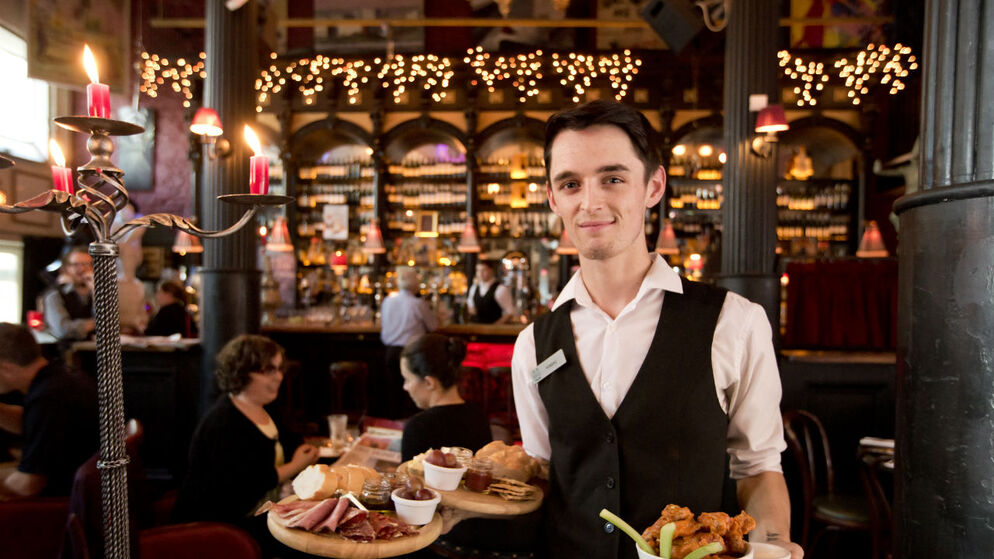 Smartly dressed waiter stands in a lively Irish pub with warm lighting, candles, and shelves of wine.