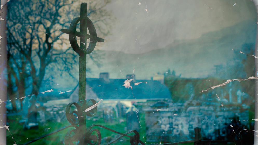 Een verweerd stenen kruis op een mistig kerkhof met kale bomen en heuvels in de verte in Ierland.