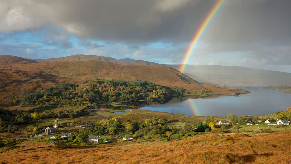 Regenbogen wölbt sich über herbstliche Hügel und einem See in Irland mit vereinzeltem Sonnenlicht und Wolken.