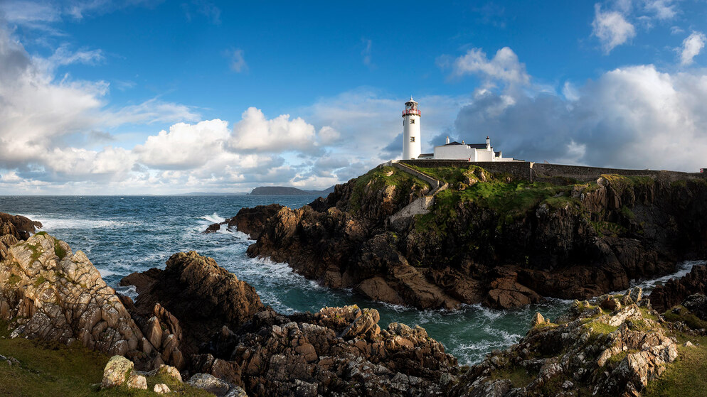 White lighthouse at Fanad Head stands above crashing waves and rugged cliffs under a bright blue sky.