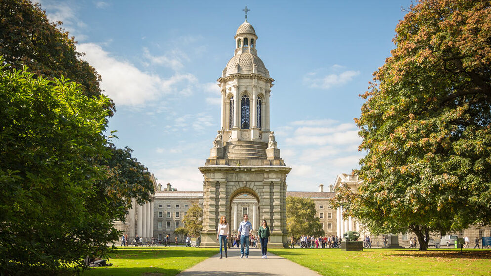 Torre del campanario del Trinity College de Dublín enmarcada por árboles con visitantes caminando por el arco.