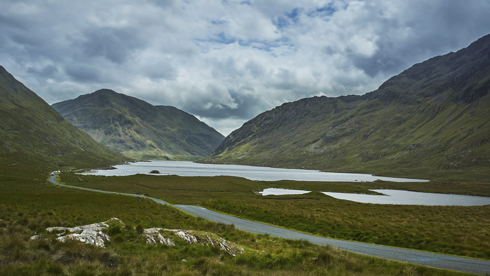 View of winding road through green mountains in Connemara, County Galway under cloudy sky.