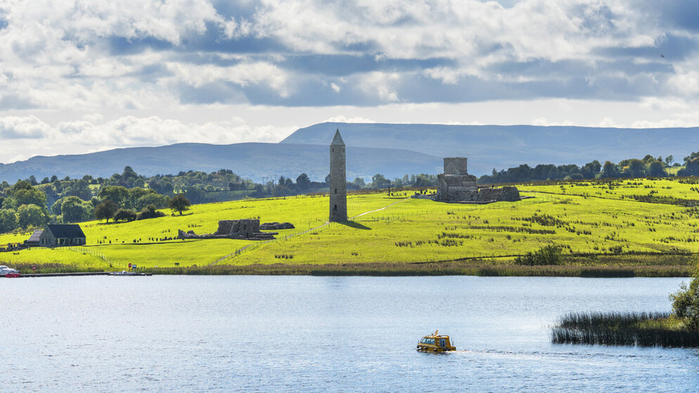 Panoramic view of Devenish Island on Lough Erne, County Fermanagh, with a round tower and church ruins framed by trees.